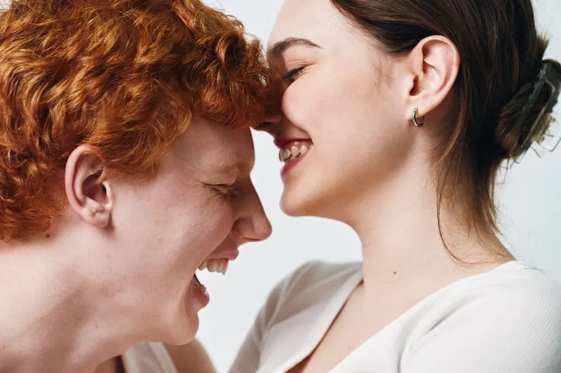 Young couple with closed eyes smiling and touching foreheads in white clothes on light background. Emotional connection, affection, love, happiness, togetherness and intimacy.