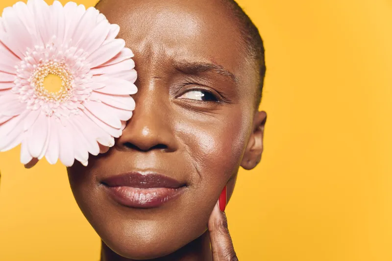Close-up of a young black woman with dark skin holding a pink gerbera flower near her face. She looks thoughtful against a bright yellow background. Concept of natural beauty and skincare.