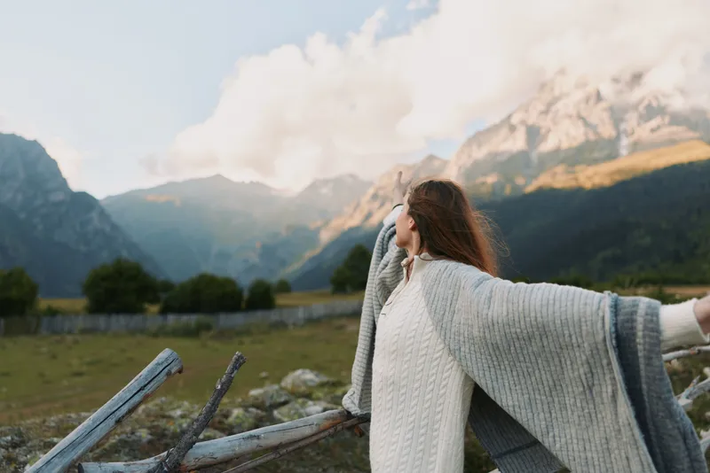 Woman in a chunky knit shawl stands with outstretched arms in a mountain meadow, embracing outdoor lifestyle and serene natural beauty.