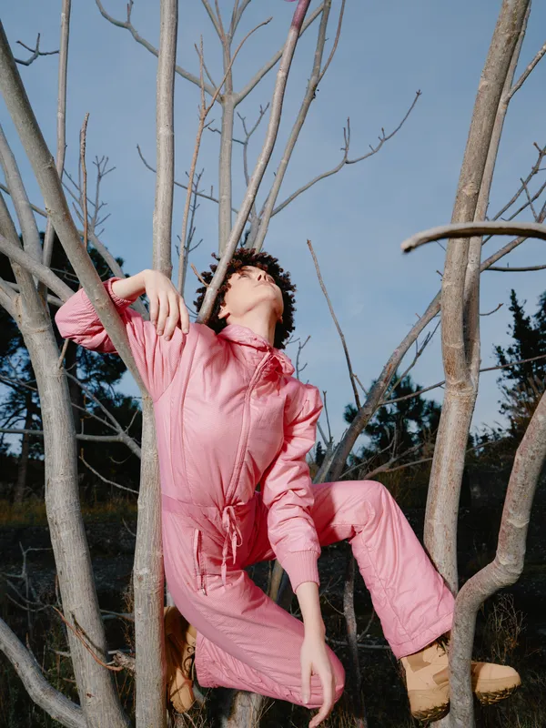 A model in a pink jumpsuit poses dramatically among leafless tree branches, looking upward. Creative fashion photography with bold composition and natural outdoor lighting.