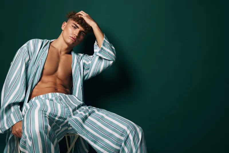 Young man with curly hair wearing striped pajamas sitting on stool against dark green background. Casual relaxed pose with hand on head and bare chest visible.