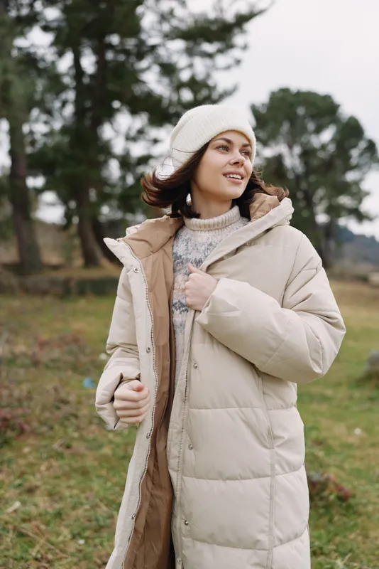 Woman in a beige puffer coat outdoors, walking in a park on a chilly day, cozy knit sweater and beanie, soft natural light against autumn scenery and a gentle breeze.
