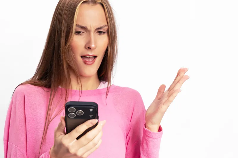 Young woman in pink sweater looking surprised and confused at smartphone screen, isolated on white background. Concept for unexpected news, disbelief, and mobile communication.