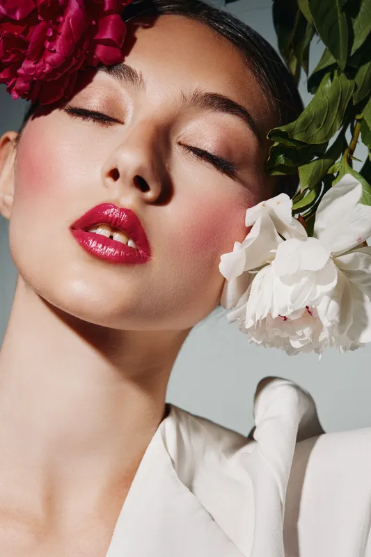 Close-up portrait of woman with bright red lips and glossy eye makeup wearing flowers in her hair and white fabric over shoulder. Concept for beauty, fashion, and creative style.
