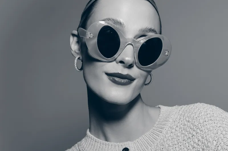 Stylish young Caucasian woman with fair skin wears large round sunglasses and hoop earrings, posing confidently in studio.