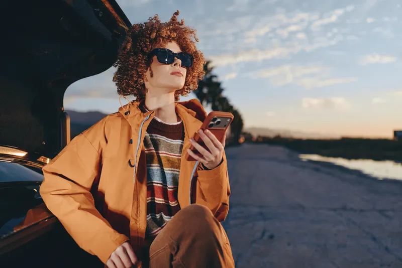 Woman lifestyle outdoors at sunset by the ocean, wearing sunglasses and an orange jacket, curly hair, seated near a car, holding a smartphone, relaxed smile, warm sky golden evening breeze.