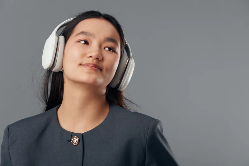 Woman wearing headphones and gray jacket standing against gray background looking to side with calm expression. Portrait of young female enjoying music or audio in studio setting.