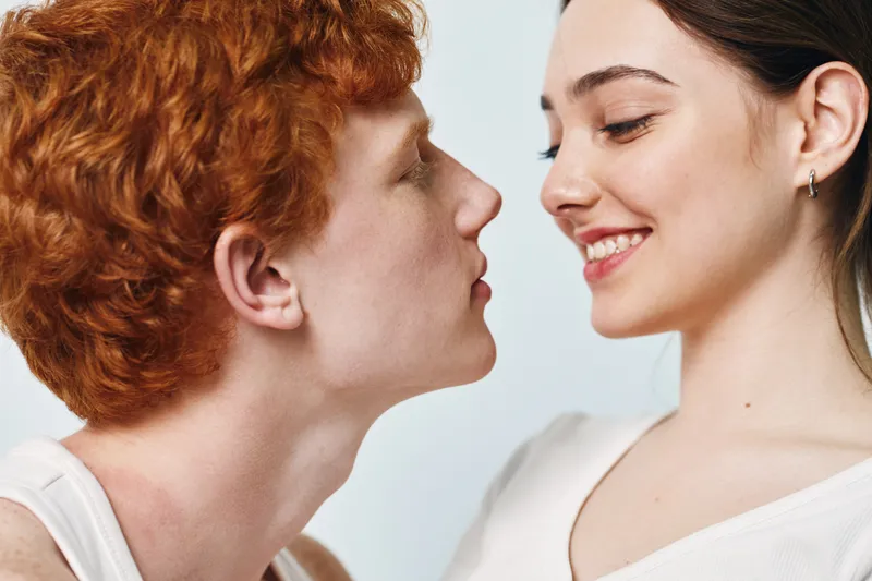 Couple, young man and woman in white tops smiling and looking at each other closely on light background. Facial expressions, intimacy and affection in studio portrait.