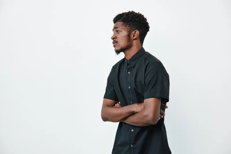 Young black man with dark skin wearing black shirt standing with arms crossed looking sideways on white background. Male model with afro hairstyle and casual clothing in studio.