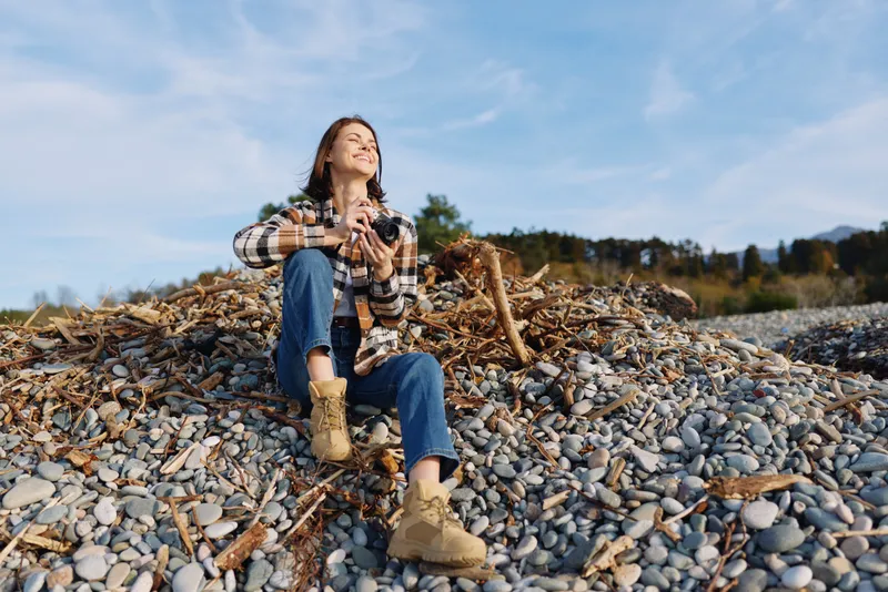 Woman outdoor sitting rocks in plaid top and boots, holding camera, enjoying nature under clear sky. A relaxed lifestyle portrait on a rocky shore. The scene emphasizes calm, adventure, and outdoor