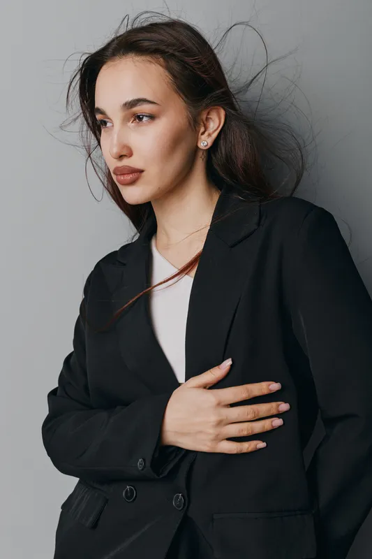 Young woman with long hair wearing black blazer and white shirt posing with hand on stomach against gray background in studio. Serious expression and natural makeup with earrings.
