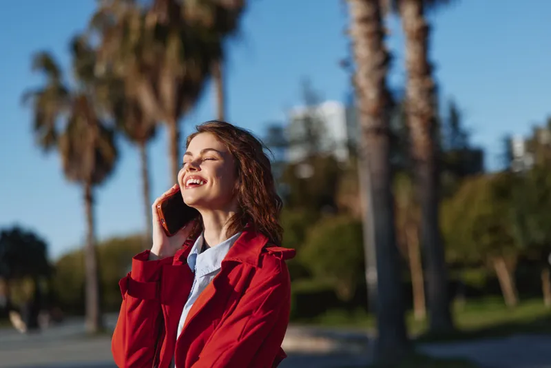 A young woman in a red jacket smiles happily while talking on a smartphone outdoors with palm trees and blue sky in the background on a sunny day.