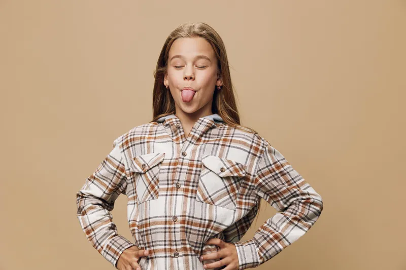 Girl in plaid shirt standing with hands on hips and sticking tongue out with closed eyes on beige background. Portrait of playful child showing funny face expression.