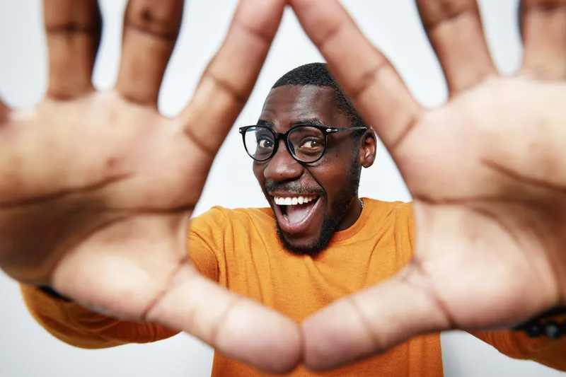 African American man with dark skin and glasses smiling and showing hands close to camera in casual orange sweater on white background.