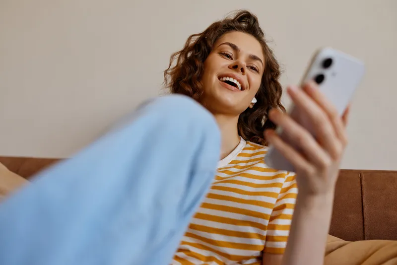 Cheerful woman relaxing at home on a couch, portrait, smiling while using a smartphone, casual lifestyle indoors, cozy living space, candid moment with relaxed posture barefoot glow cozy.