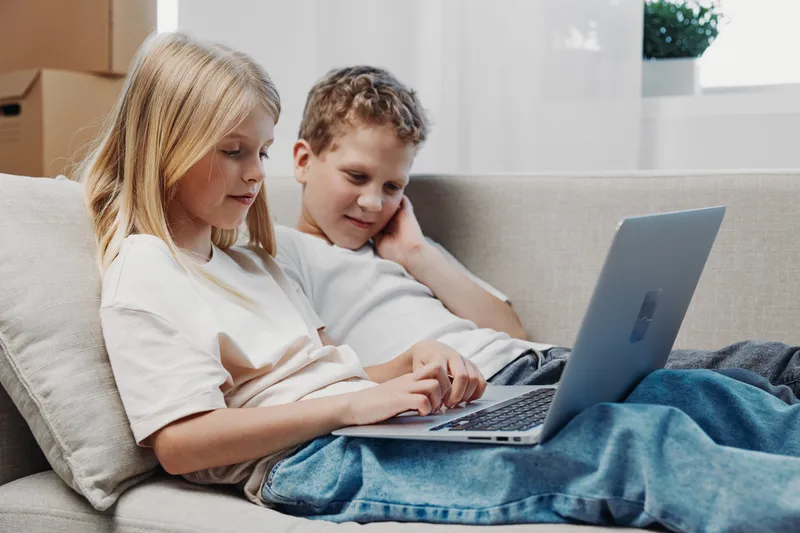 Two children sitting on a sofa using a laptop together in a bright living room. Casual home atmosphere with natural light and relaxed mood.