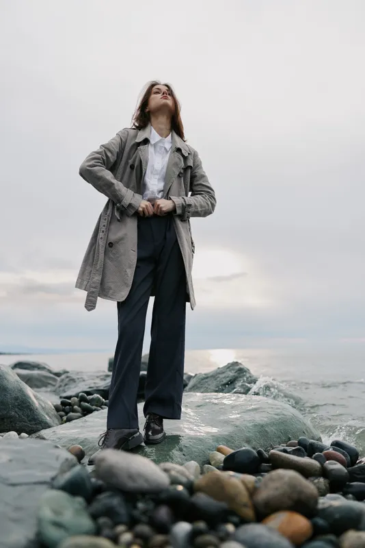 Confident woman wearing long coat and wide pants stands on rocky shore near ocean under cloudy sky. Concept of fashion, autumn style, and outdoor lifestyle with natural coastal environment.