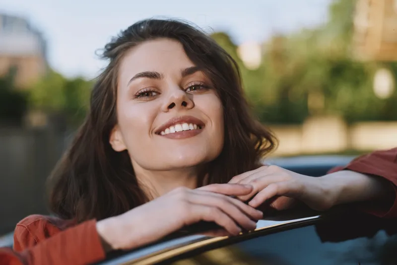 Smiling woman with brunette hair leans on a car window, enjoying a sunny day and casual conversation. In a soft outdoor background with warm light, and a breeze