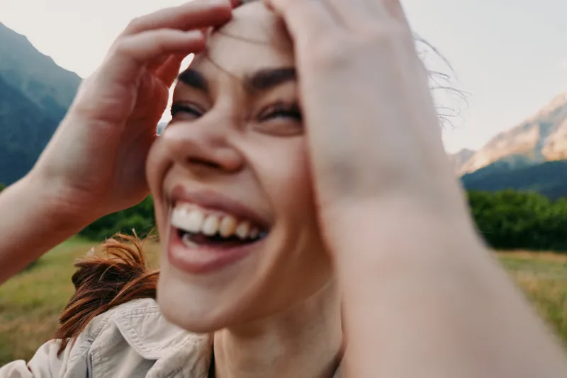 A candid outdoors portrait of a woman laughing in nature, with mountains in the background, conveying joy, freedom, and a carefree lifestyle.
