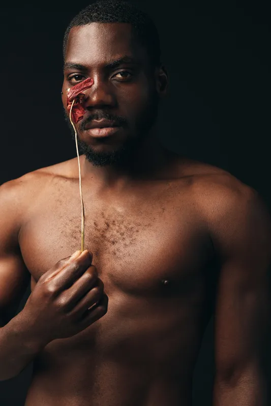 Black man with bare chest holding dried flower near face in creative conceptual visual style. Portrait of African ethnicity with artistic expression on dark background for modern art.
