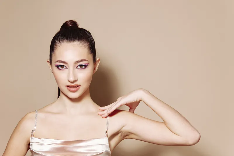 Young woman with elegant updo hairstyle and bold pink eye makeup poses gracefully wearing a satin dress on beige background. Beauty and fashion creative portrait concept.