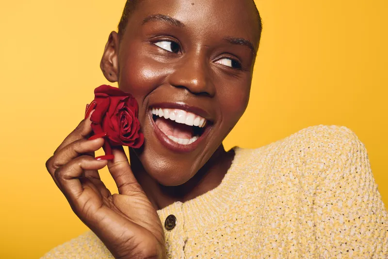 Happy African woman with dark skin holds red rose near face smiling brightly on yellow background. Concept of beauty, joy, love, skincare, and natural happiness with floral element.