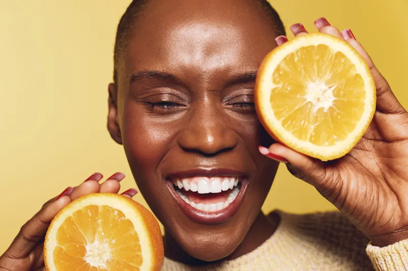 Happy African woman with dark skin holds two orange halves near face smiling with closed eyes on yellow background. Concept of fresh fruit, healthy lifestyle and natural beauty.