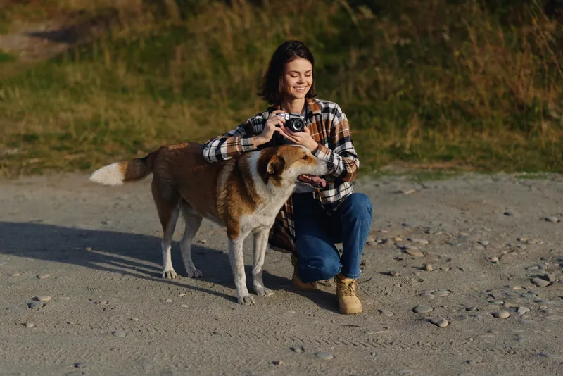 Dog and woman on a sandy beach, outdoor photography moment as she holds a camera and smiles, capturing a candid portrait and natural companionship in golden light.