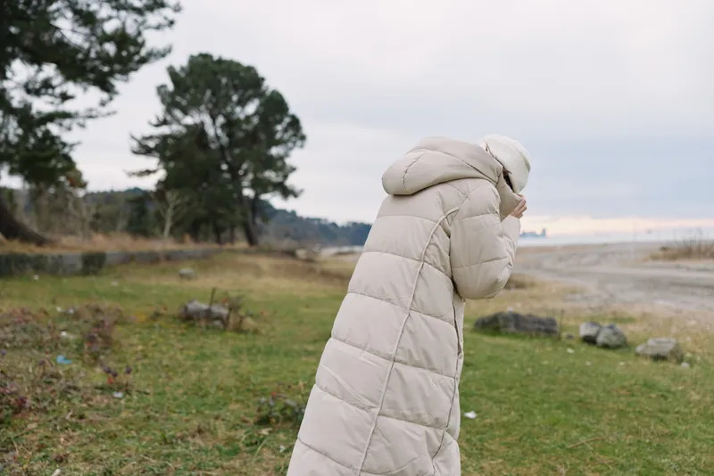Person wearing a long beige puffer coat walks along a windy shoreline, with grass, trees, and a calm cloudy sky in the background.