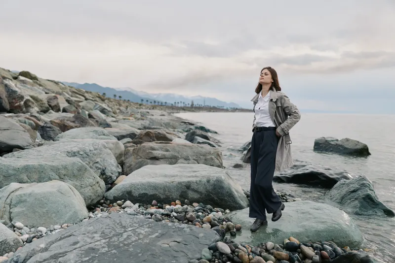 Young woman stands on rocky coast near calm sea under cloudy sky. She wears casual clothes and looks relaxed, enjoying nature and peaceful outdoor scenery by the water.