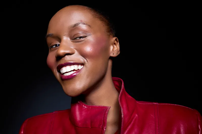 Close-up of a smiling African woman with dark skin wearing a red leather jacket on black background. Portrait of confident black female model with natural beauty and joyful expression.