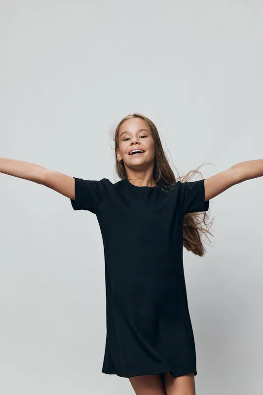 Girl in black dress with long hair smiling and spreading arms wide for happiness and freedom against plain gray background in studio portrait.