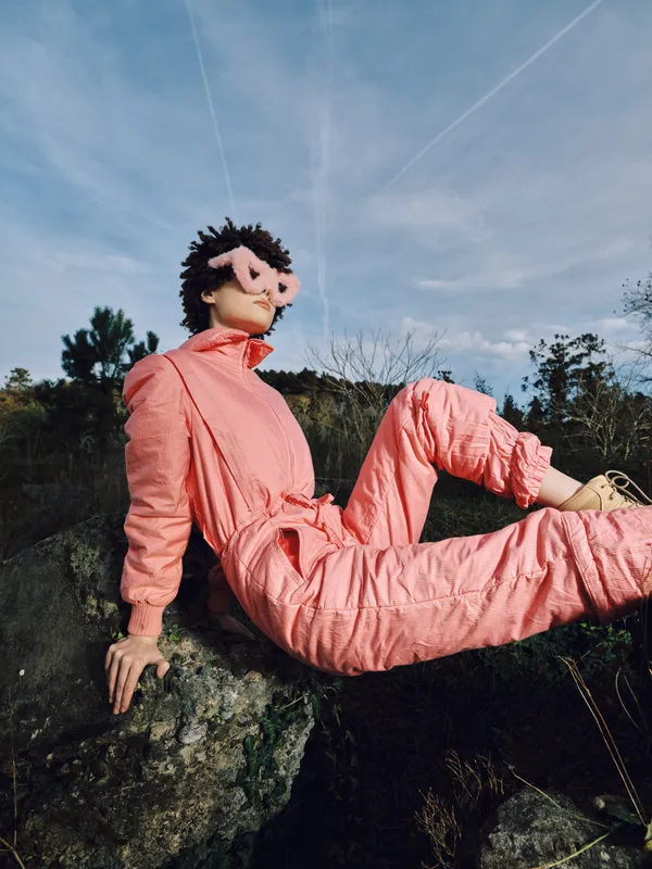 Puppet figure in a pink suit sits on a rocky outcrop outdoors, wearing a mask and a relaxed pose under a clear blue sky among sparse trees and shrubs