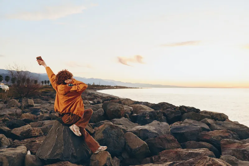 Woman lifestyle seaside sunset, casual outfit, sits on rocks, raises arms and dances, joyful smile, outdoors on shoreline, calm ocean and soft sky, warm light, breeze