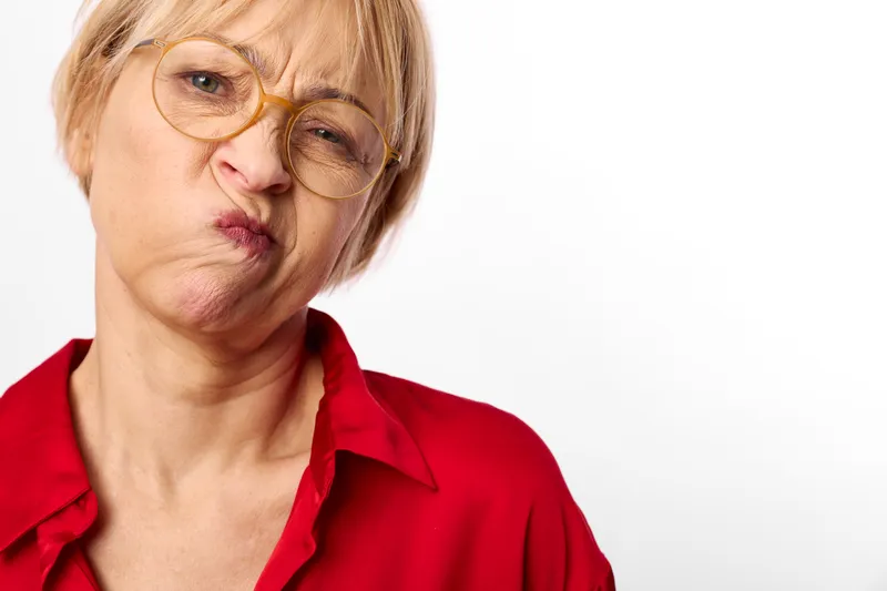 Senior woman portrait closeup with glasses, red shirt, skeptical expression, studio lighting, isolated on white background, soft skin texture, calm still tension
