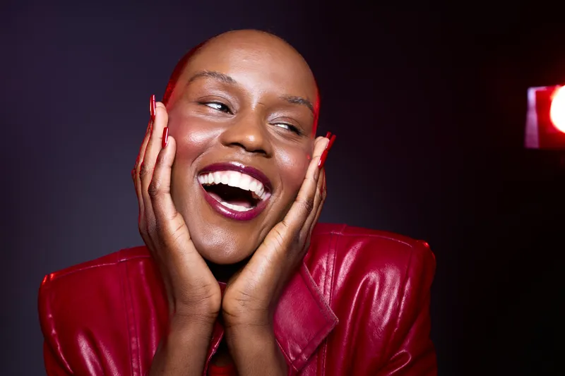 Smiling black woman with dark skin wearing a red leather jacket expresses joy with hands on face. Studio portrait with dramatic lighting and vibrant mood. Concept of happiness and confidence.