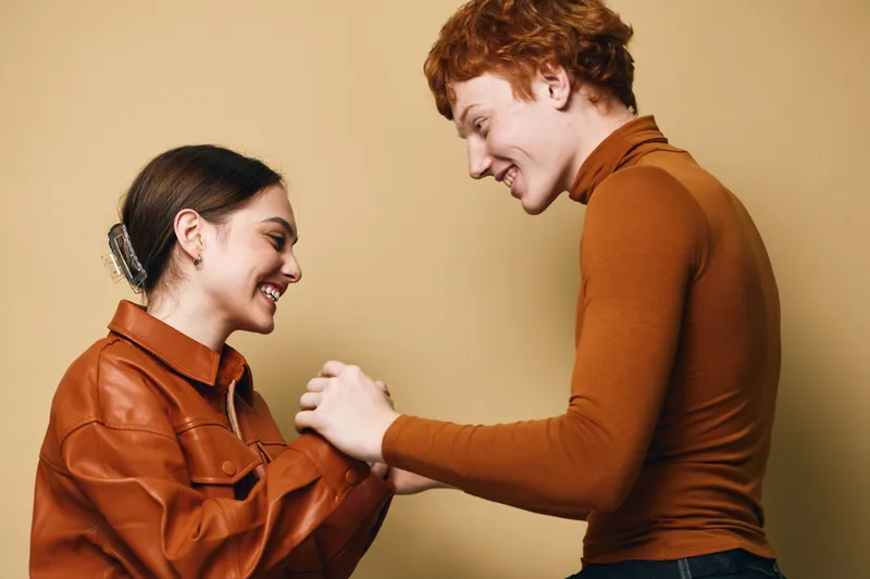 Couple, young man and woman smiling and holding hands in casual clothing indoor with warm background. Positive relationship moment with friendship and connection between two people.