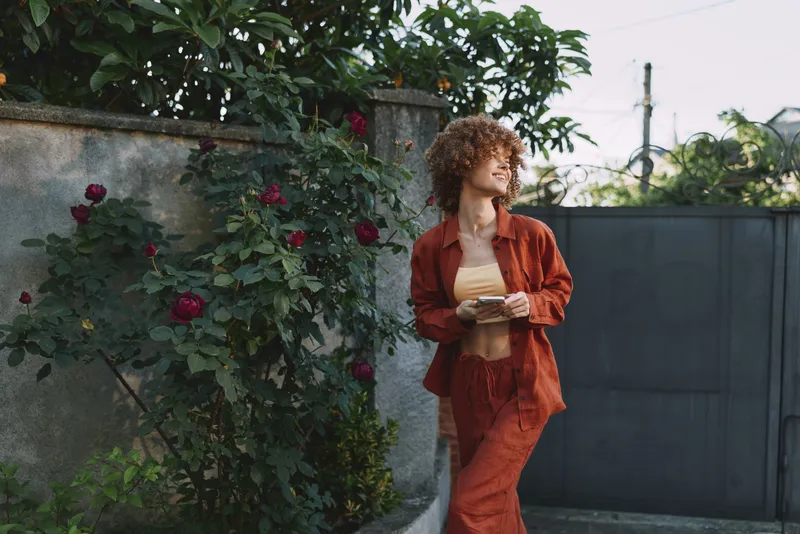 Young woman with curly hair in a rust suit walks along a garden path beside blooming roses, a warm confident mood on a sunlit day, smiling softly