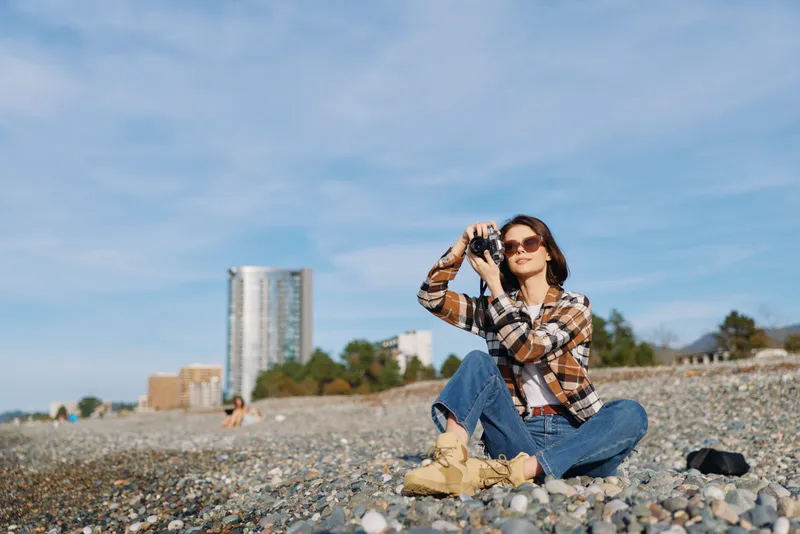 Woman photographer sits on a pebble beach with a camera raised, wearing a plaid shirt, jeans and boots. Bright sky, distant buildings, and a calm seaside moment captured for outdoor lifestyle and