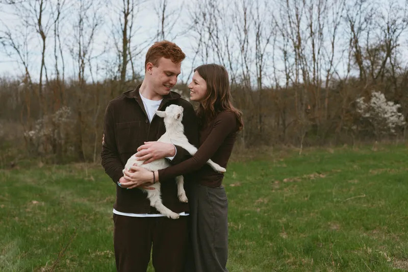 Cheerful couple outdoors, holding a playful goat kid, warm interaction, sunlit meadow, relaxed posture, natural colors, candid moment, gentle breeze, rustic charm