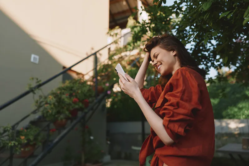 Young woman in a red blouse chats on her phone outdoors in a sunny courtyard, smiling softly as warm light filters through trees and a quiet space around her