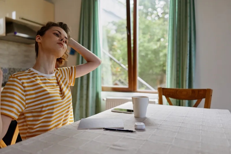Relaxed woman at home sits at a dining table with a notebook and mug, bathed in natural light from the window, wearing a striped shirt, capturing everyday life and a calm morning routine