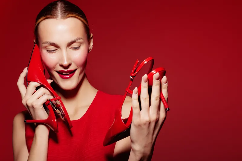 Young woman in red dress holds red high heel shoes near face with closed eyes and smile. Bright red background creates vibrant mood. Concept of fashion, style, and elegance.