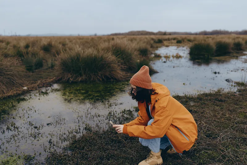 Woman outdoor lifestyle in nature, wearing raincoat and hat, standing by pond in wetland, adjusting jacket while walking through muddy grass, cloudy daylight, gentle wind, natural colors