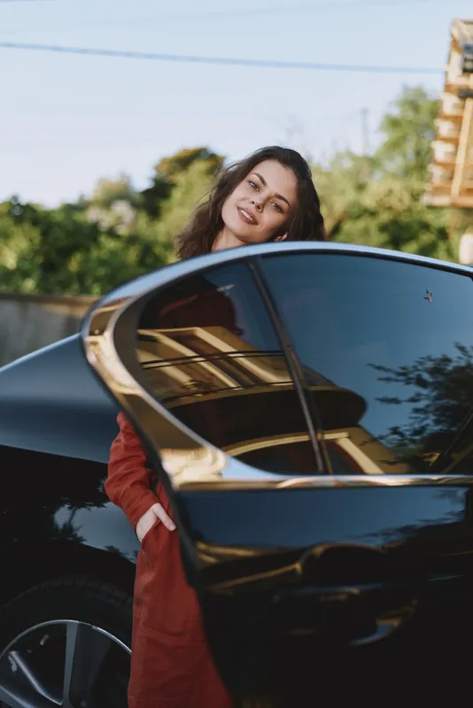 Woman lifestyle portrait beside a sleek car, smiling toward the camera in an outdoor urban setting, casual everyday mood and relaxed vibe, candid and friendly