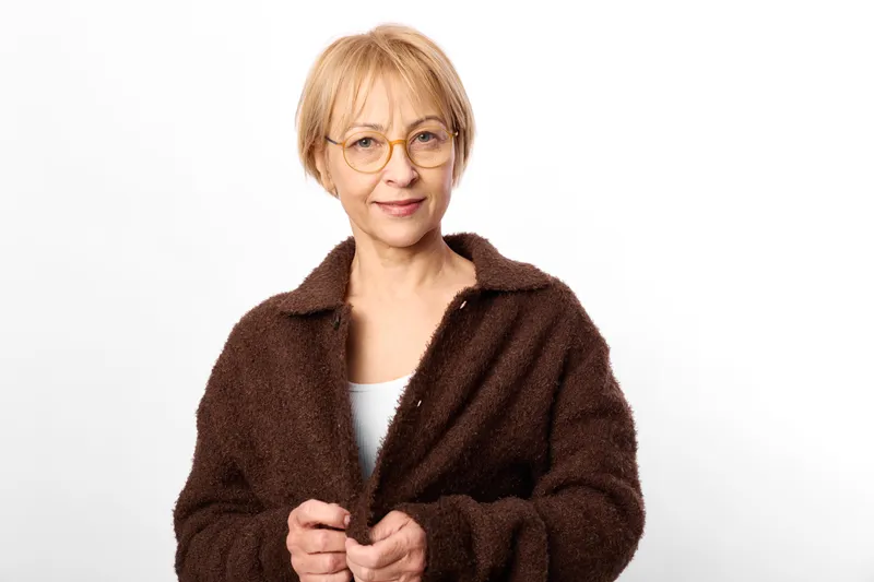 Woman portrait, glasses, elderly mature face, wearing a dark cardigan, indoor studio background, smiling gently, relaxed posture, soft light, clean minimal copyspace for text here