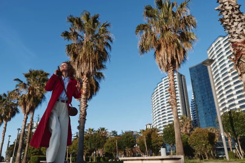 A woman in a red coat stands beneath tall palm trees in a sunny city park, looking up while talking on her phone, delivering a confident urban fashion moment against modern high rises.