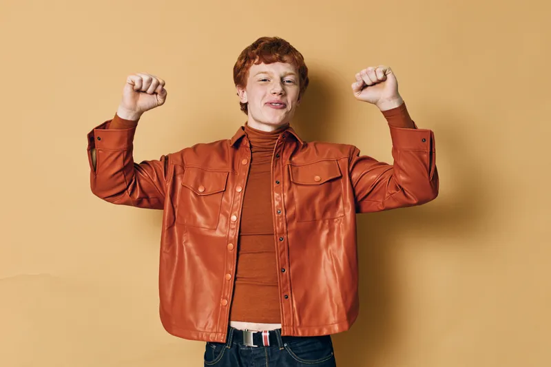 Young man with red hair wearing a brown shirt and orange jacket, smiling confidently with raised fists against beige background, showing excitement and positive emotion.