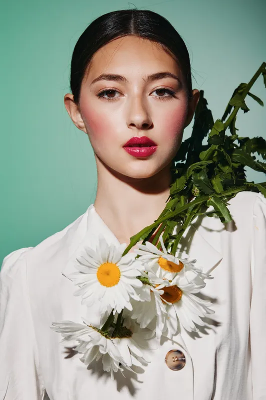 Elegant woman with bold red lipstick and flawless makeup poses with white daisies on her shoulder against pale background. Concept for beauty creativity and fashion editorial.