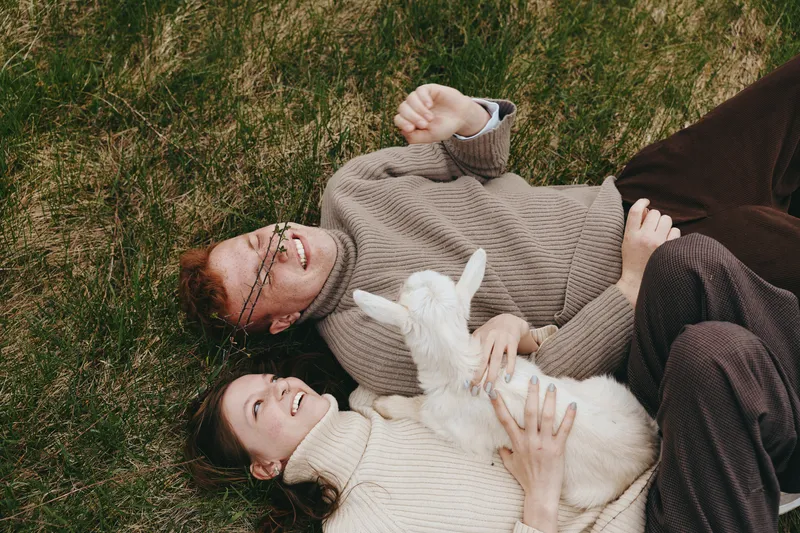 Relaxed couple outdoors on grass in knit sweaters, sharing a moment with a playful goat kid. Warm natural light enhances a cozy lifestyle scene of affection and companionship in nature.